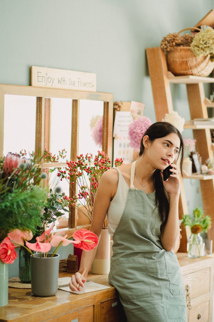 Woman florist in an apron talking on a phone surrounded by flowers in a shop.